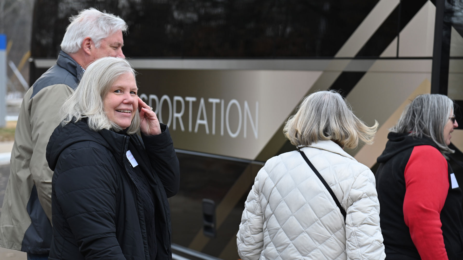 Smiling older woman with gray hair in a black jacket stands beside a bus with the partial word 'PORTATION' on its side."