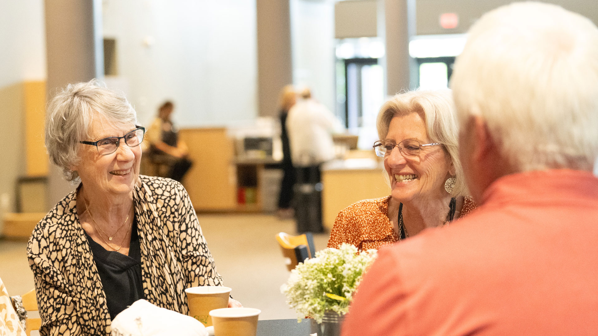 Two smiling elderly women sit at a table in a bright cafeteria, coffee cups in front of themIs this?