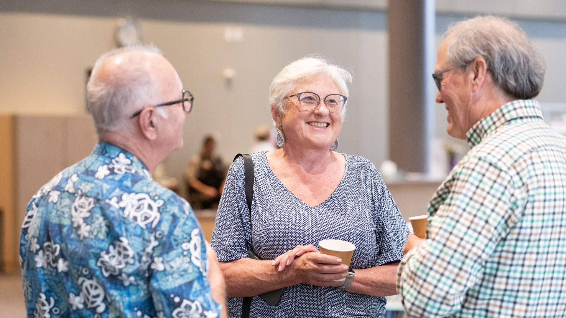 Three seniors stand in a bright room, smiling and chatting while holding disposable coffee cups.