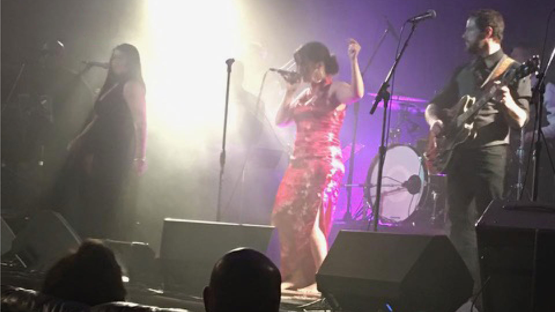Female lead singer in a red dress performing on stage, with guitarist to her right and drums in the background under bright lights.