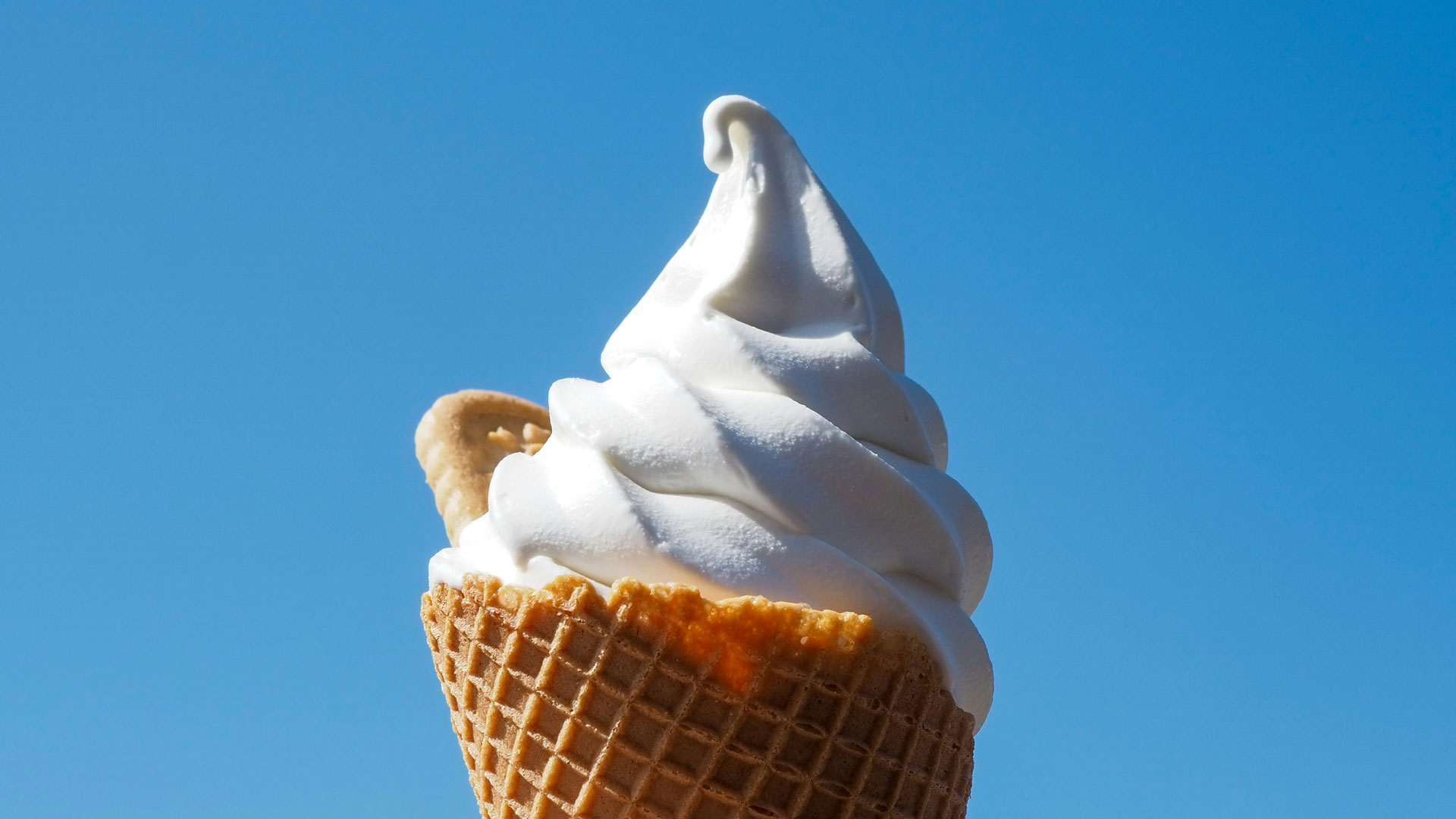 Soft-serve vanilla ice cream in a waffle cone against a clear blue sky.