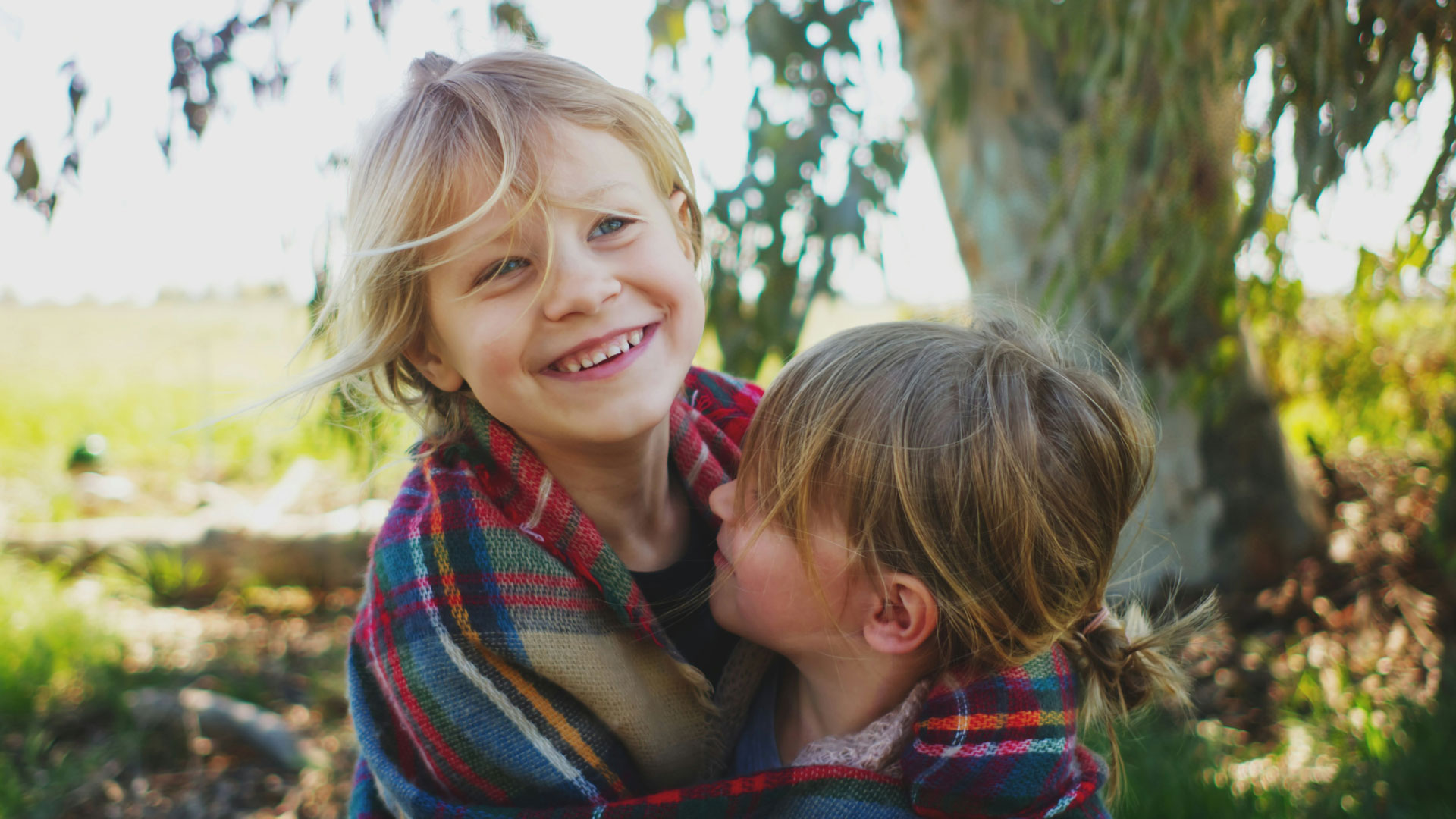 Two young children outdoors; a blonde girl smiles while hugging a friend, wrapped in a colorful plaid blanketie.