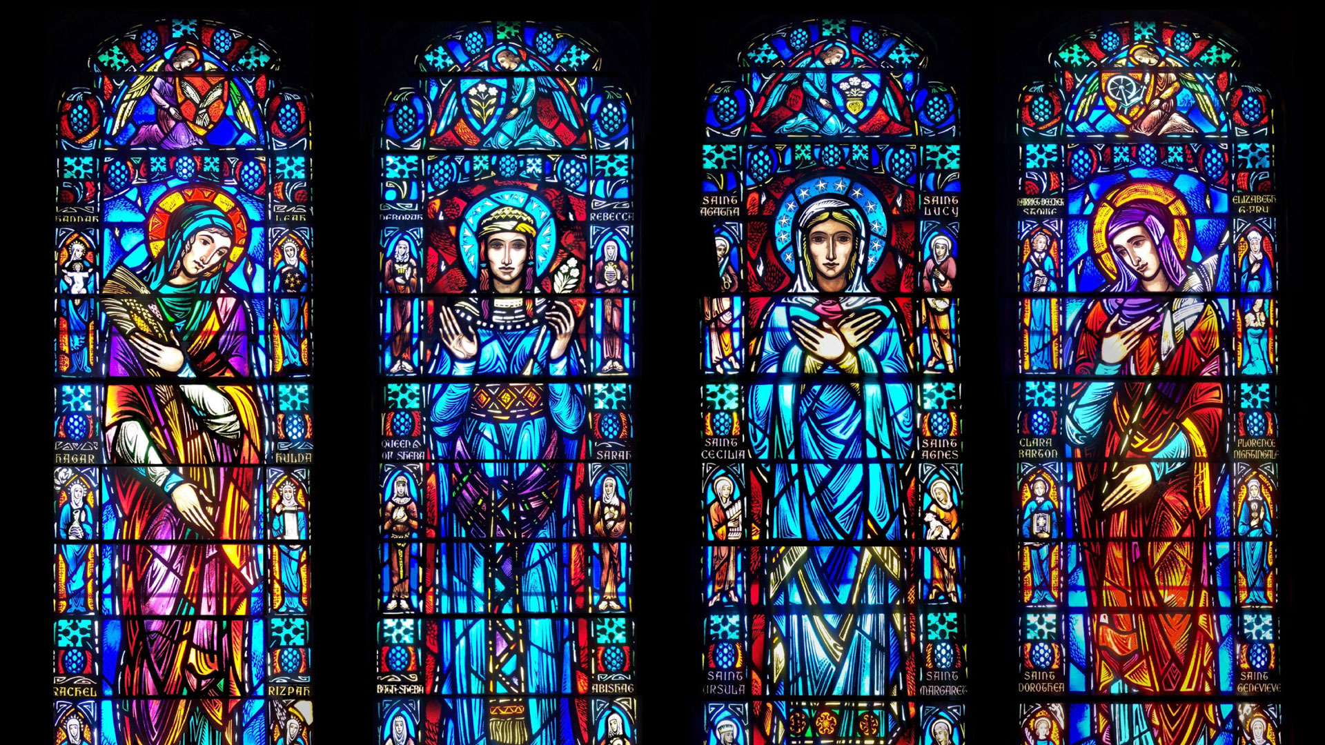 Four tall stained-glass panels in a row, depicting saints and angels with halos amid vivid blue, red, and gold patterns.