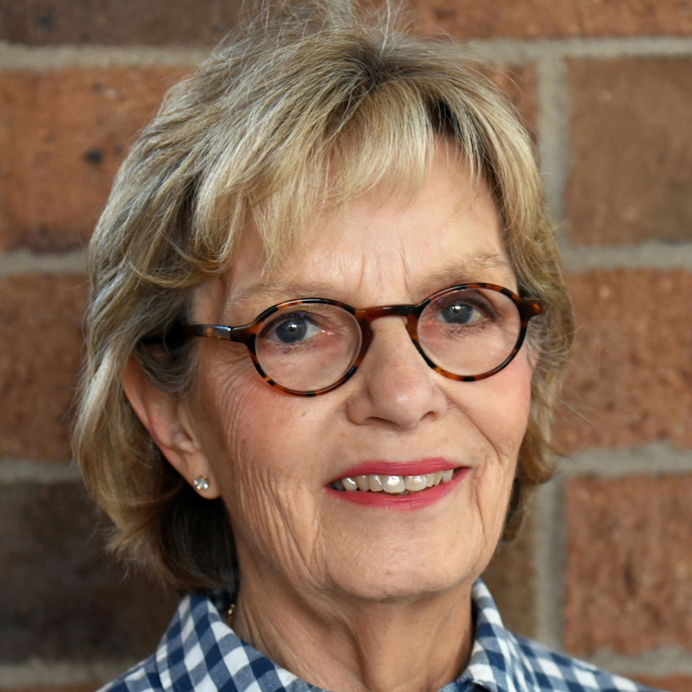 Smiling older woman with tortoiseshell glasses and a blue plaid shirt against a brick wall background