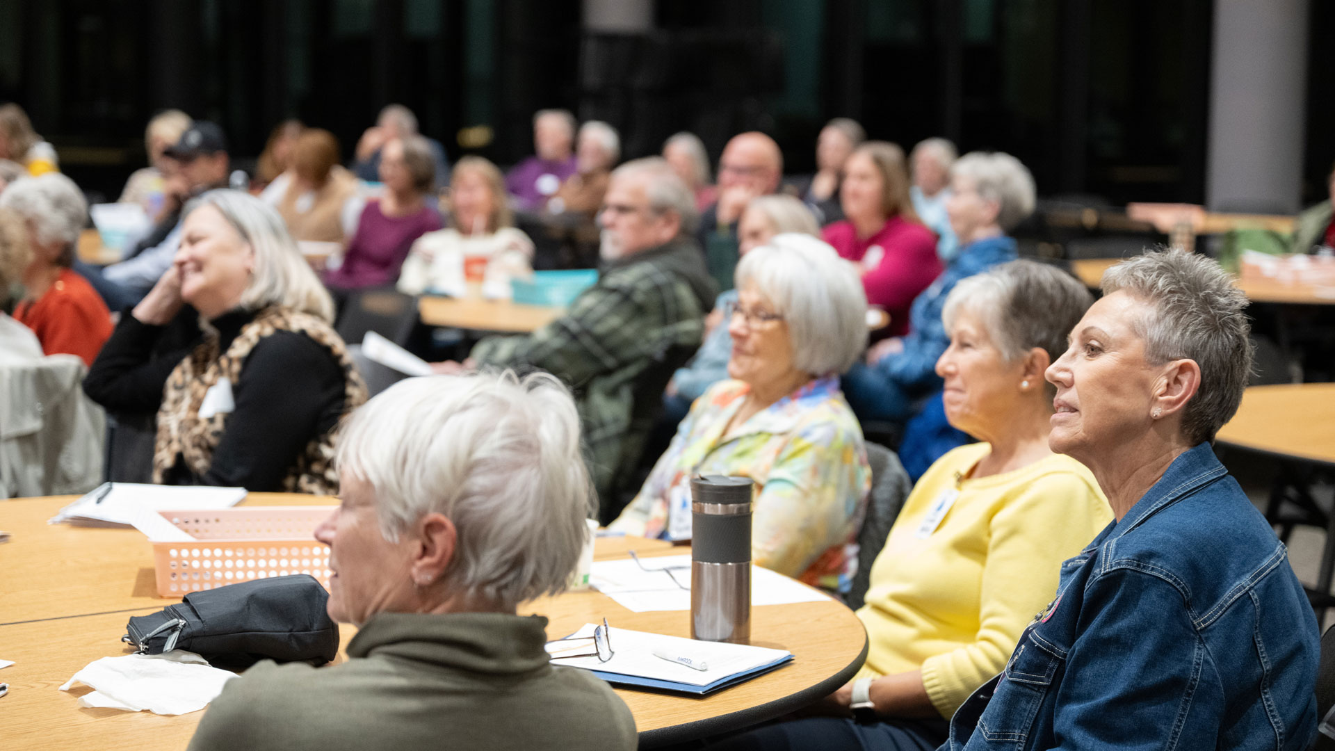 Group of older adults seated at round tables in a classroom, listening to a presentation.