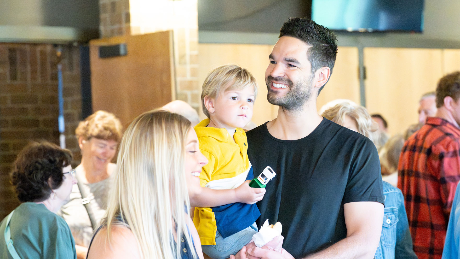 Smiling man in a black shirt holding a young boy in a yellow jacket at a crowded indoor event.