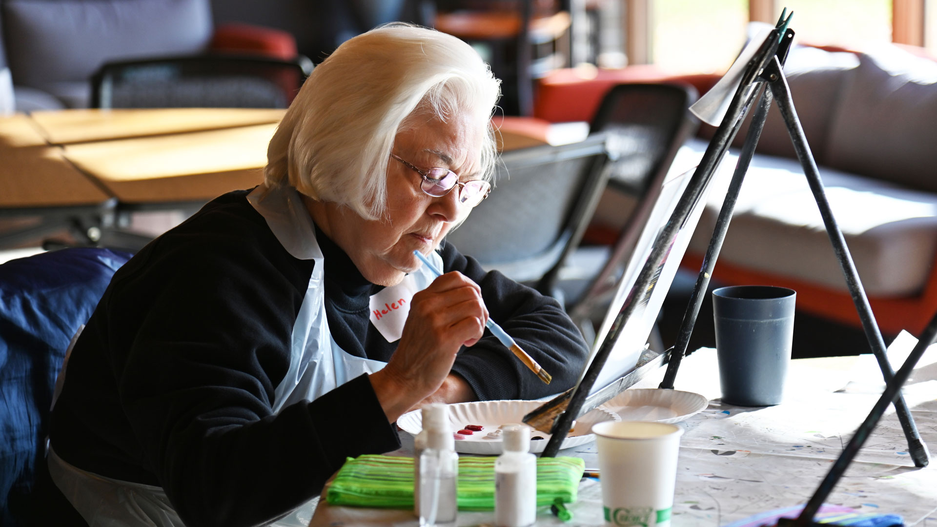 Older woman painting at a table with an easel, focused on her artwork, wearing a black sweater and name tag.