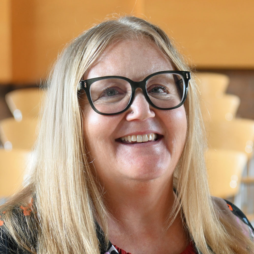 Smiling woman with long blonde hair and black glasses, indoors with wooden chairs in the background.