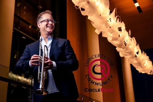 Smiling man in a dark suit holds a trumpet on stage, with a backdrop banner that reads 'Concerts for Community Circle Rock'.